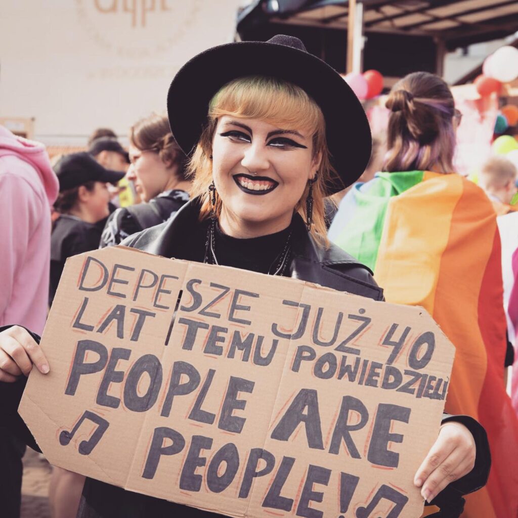 En leende person med svart hatt och svart läppstift och eyeliner håller en skylt med polsk och engelsk text: People are people! I bakgrunden syns en Prideparad.