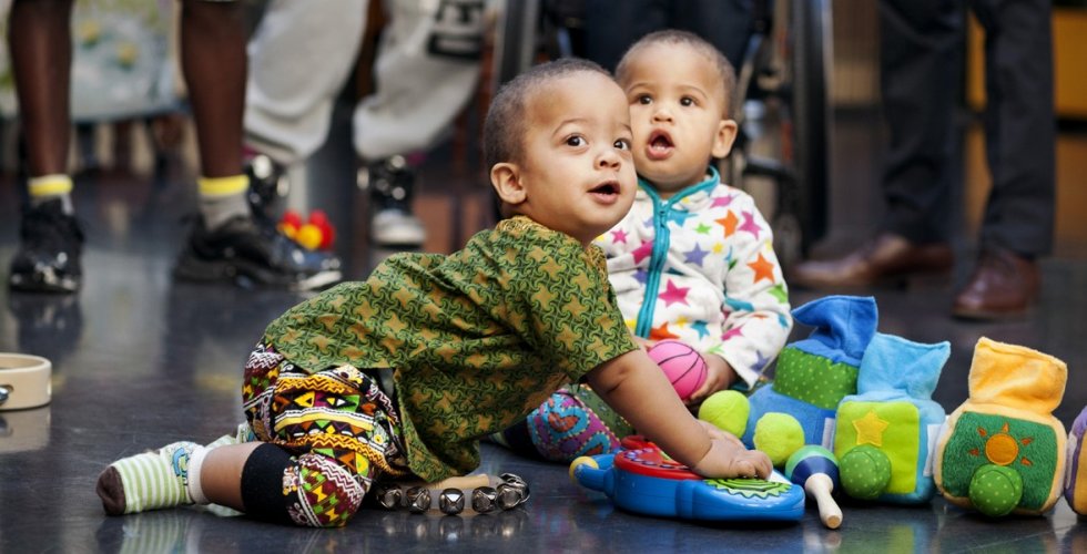 Tvillingar i ettårsåldern som leker på ett golv med fyra föräldrar i bakgrunden